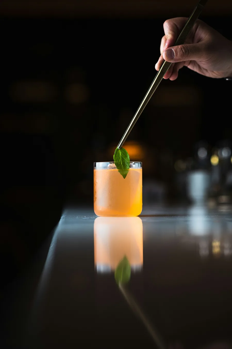 A craft cocktail being carefully garnished on a dark, reflective bar surface